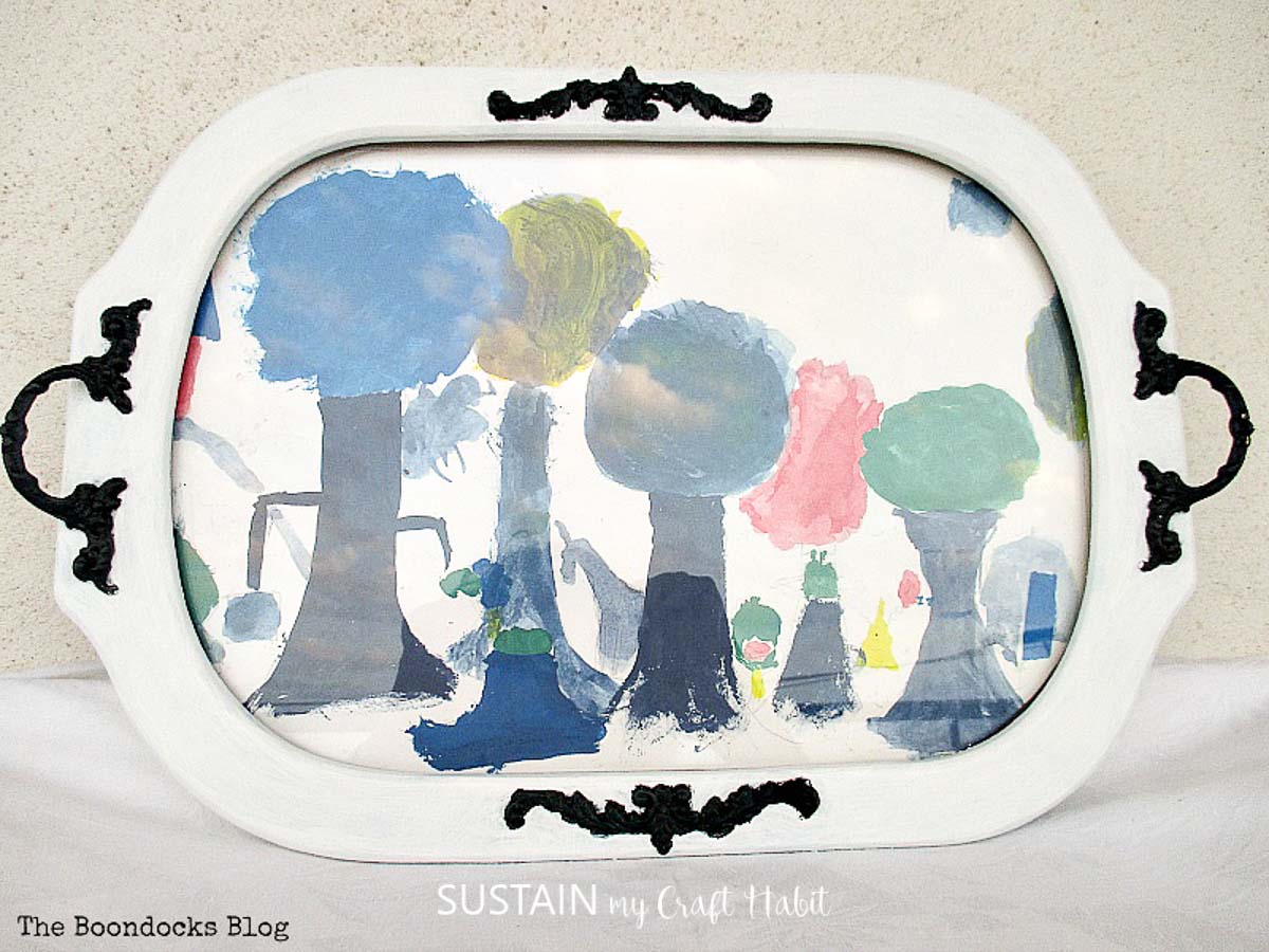 Completed upcycled serving platter with a child's painting of trees mounted behind the glass.