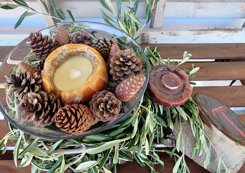 Glass bowl with another candle and pine cones, placed over olive branches, placed beside a candle and wood stump.