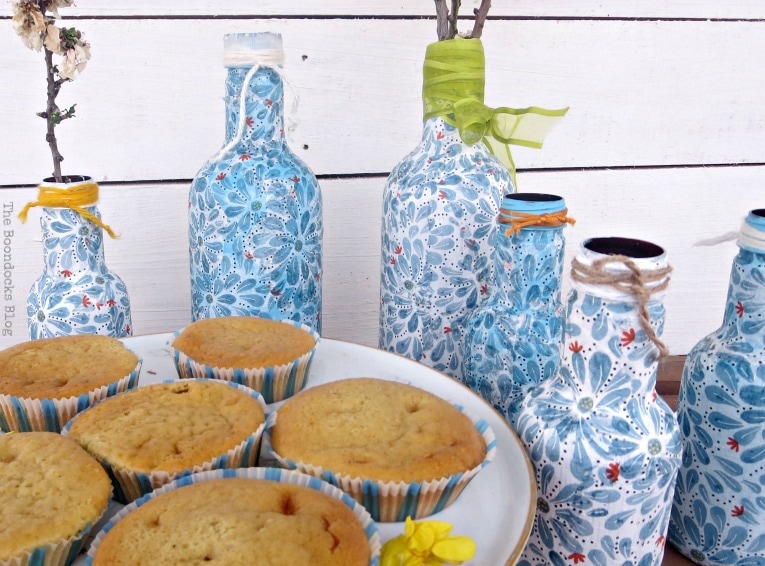 Finished decoupaged bottles next to cake stand with cupcakes.