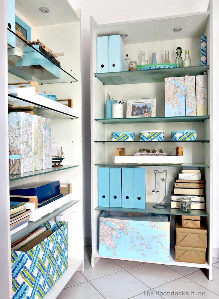 Two bookcases styled with blue colors, maps and pomegranate branches, #bookcasestyling #coastaldecor #summervignettes #upcycledmagazinefiles #upcycledcartons #mapboxes #decoupage #chalkytypepaint #organizing #storagesolutions Upcyled with paint, fabric and decoupage. Pretty and Practical Bookcases the Easy Way www.theboondocksblog.com