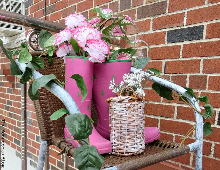 Rattan chair on which boots and wicker bottle holder are placed with flowers for an outdoor decor, #repurposeddecor #outdoordecor #curbappeal #repurposedrubberboots #fauxflowers #easyupcycleddecor #reusingbrokenchair How to re-purpose a torn rattan chair, www.theboondocksblog.com